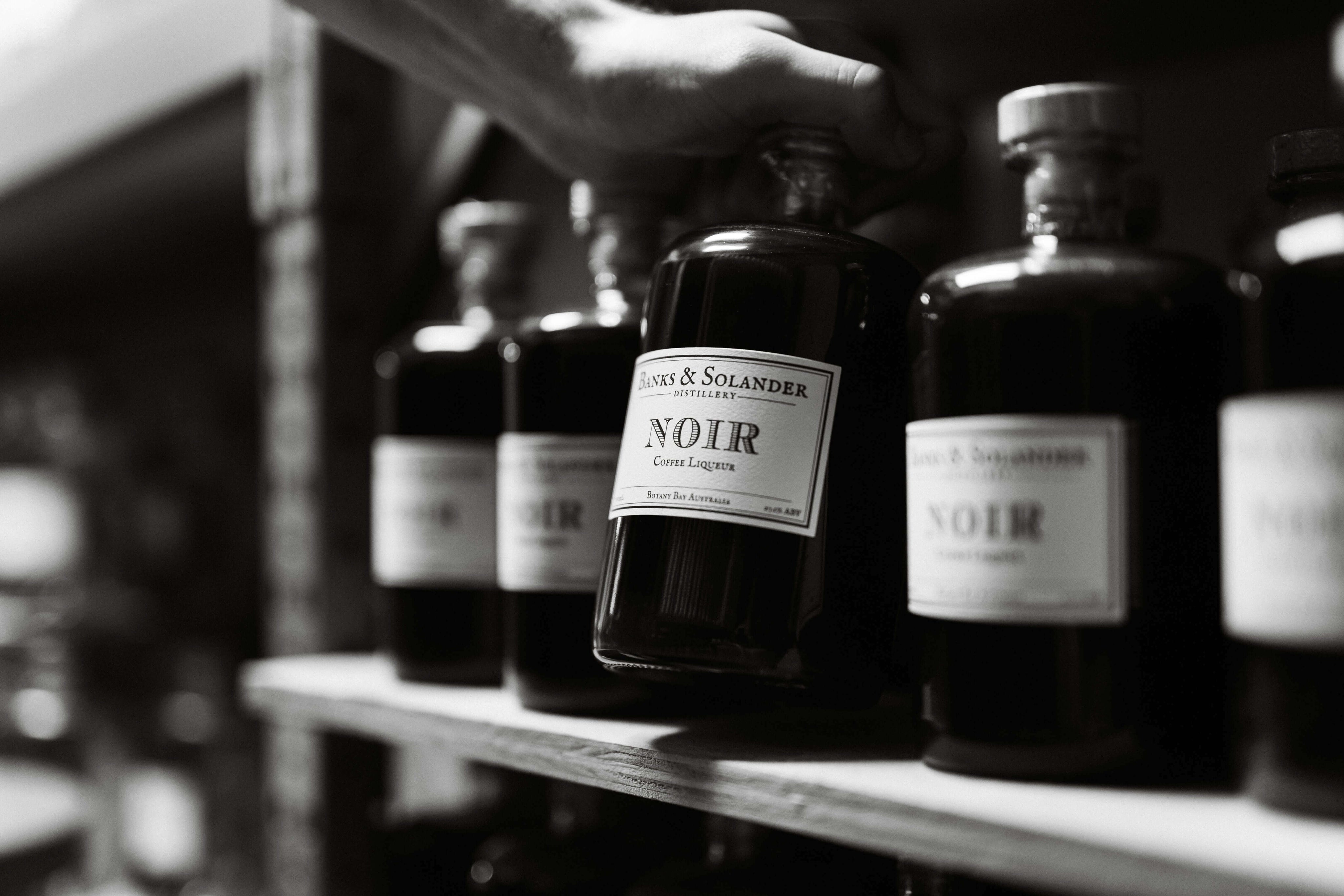 A hand reaches into a dimly lit shelf to pick up a bottle of Banks &amp; Solander Noir. The bottle has a white label with black text and sits amongst other bottles of the same drink. The image is in black and white.