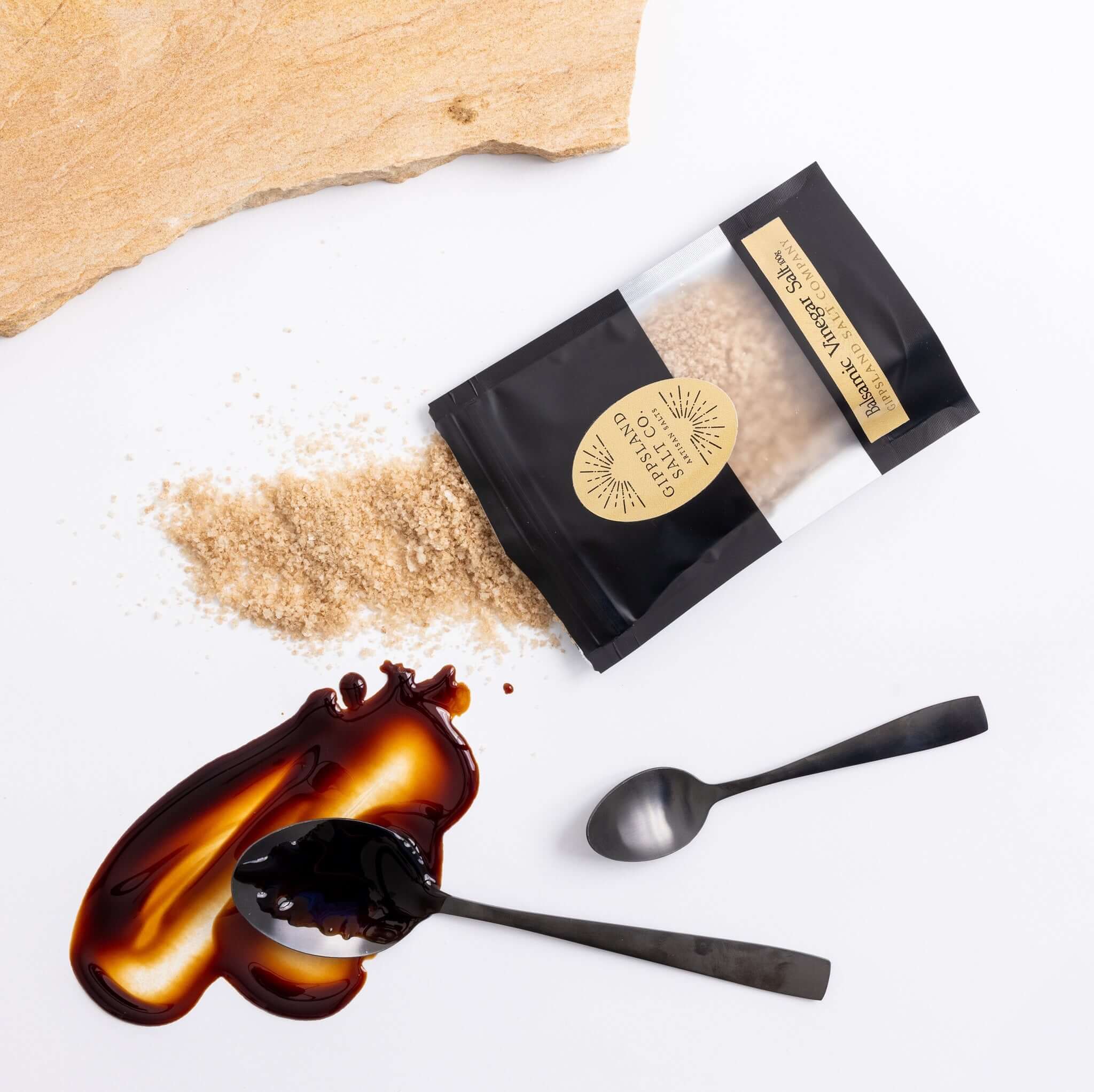 A small black pouch containing Balsamic Vinegar Salt sits on a white surface. The pouch is open, and some of the salt has spilled out. Next to it are two black spoons, one with a dark liquid spilled on it. The background includes a piece of wood and a whi