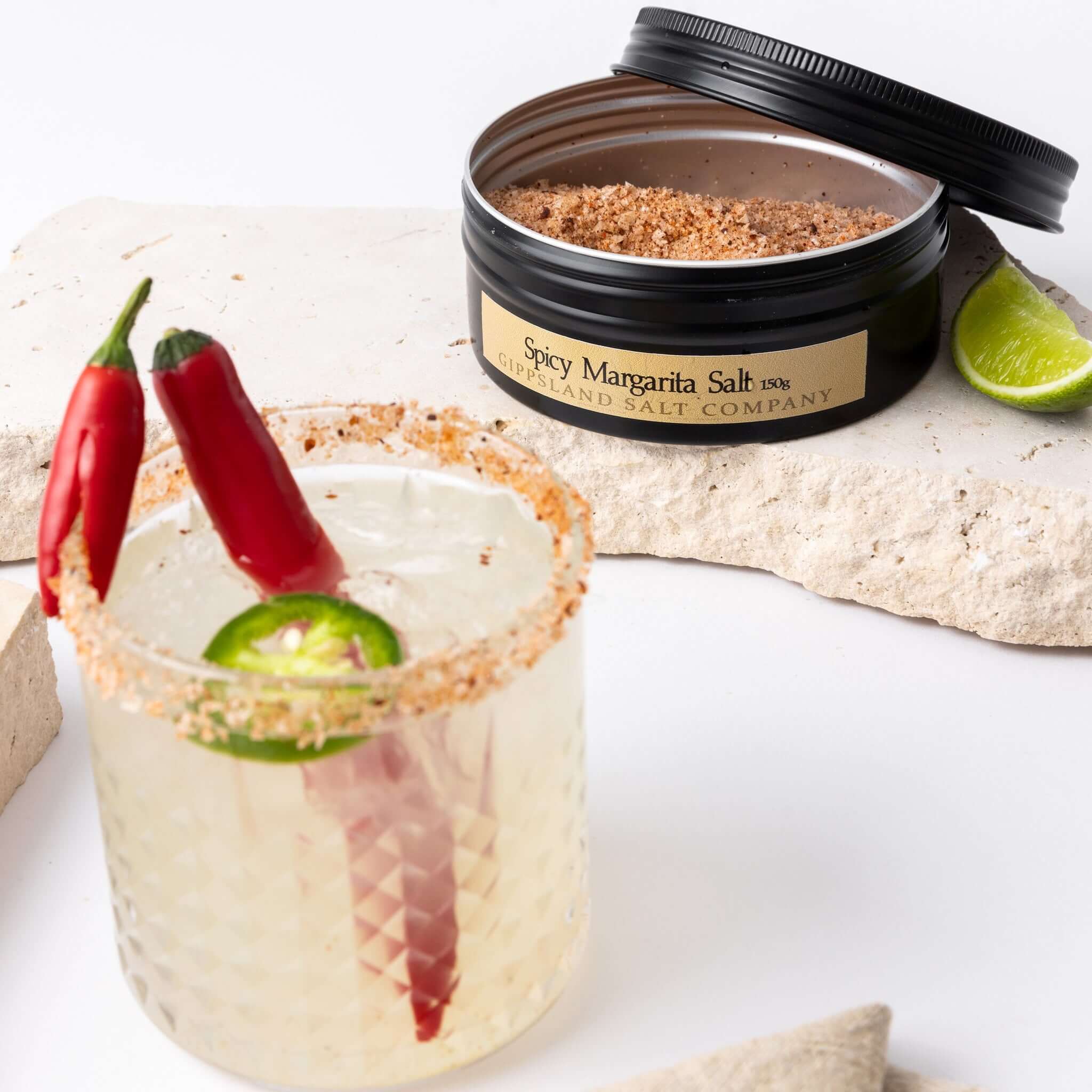 A frosty margarita cocktail with a spicy rim sits alongside an open tin of Gippsland Salt Company's Spicy Margarita Salt. Red chili peppers and a lime wedge add a vibrant touch to the scene. The image evokes a refreshing and flavorful cocktail experience.