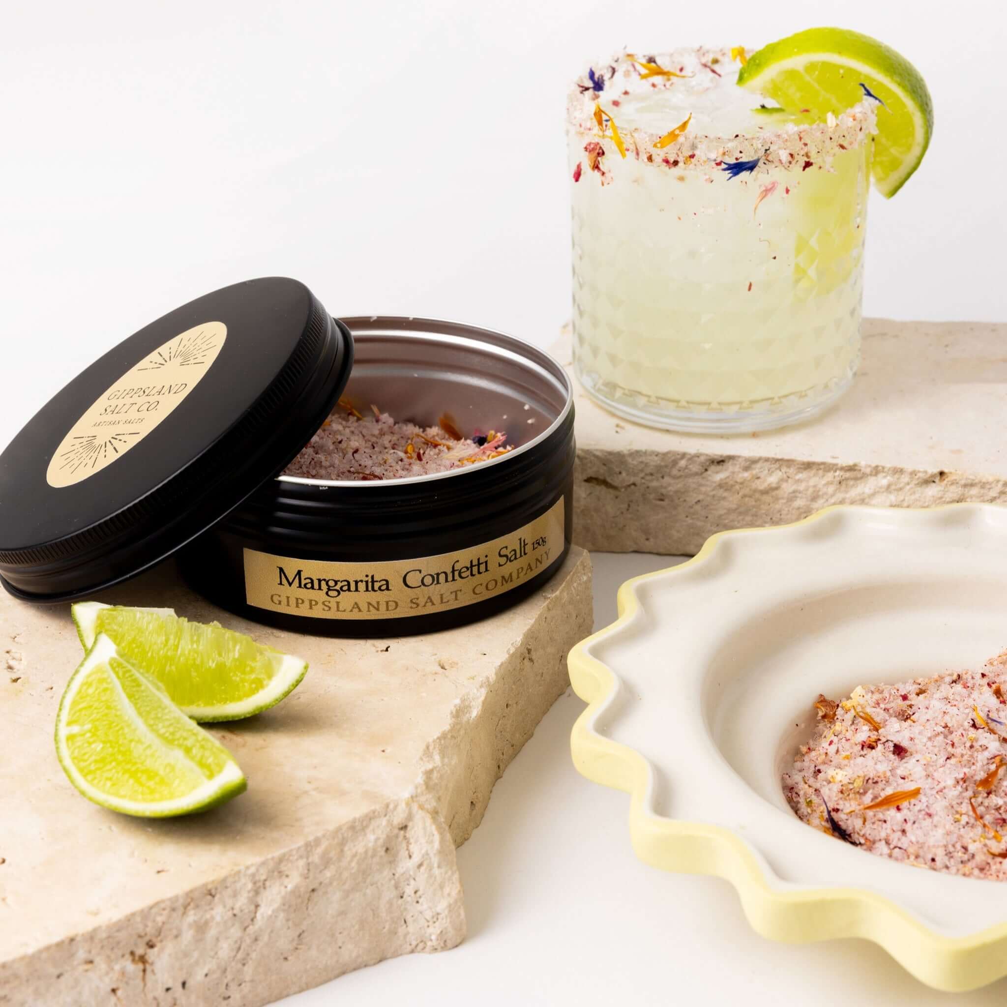 A glass of margarita with a colorful salt rim sits next to an open tin of "Margarita Confetti Salt" from Gippsland Salt Company. Slices of lime and a small bowl of the salt are also present on a textured surface. The image evokes a refreshing summer cockt
