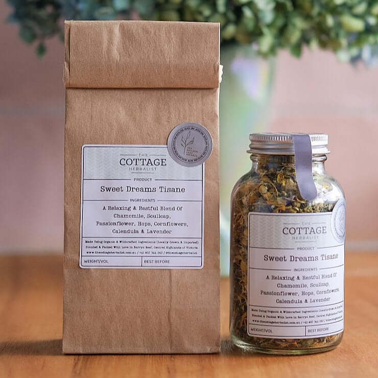 Two glass jars filled with dried herbs sit on a wooden table. The labels read "The Cottage Herbalist Y.E.P Tisane" and list the ingredients as a cooling blend of wildcrafted yarrow and elderflower with peppermint.