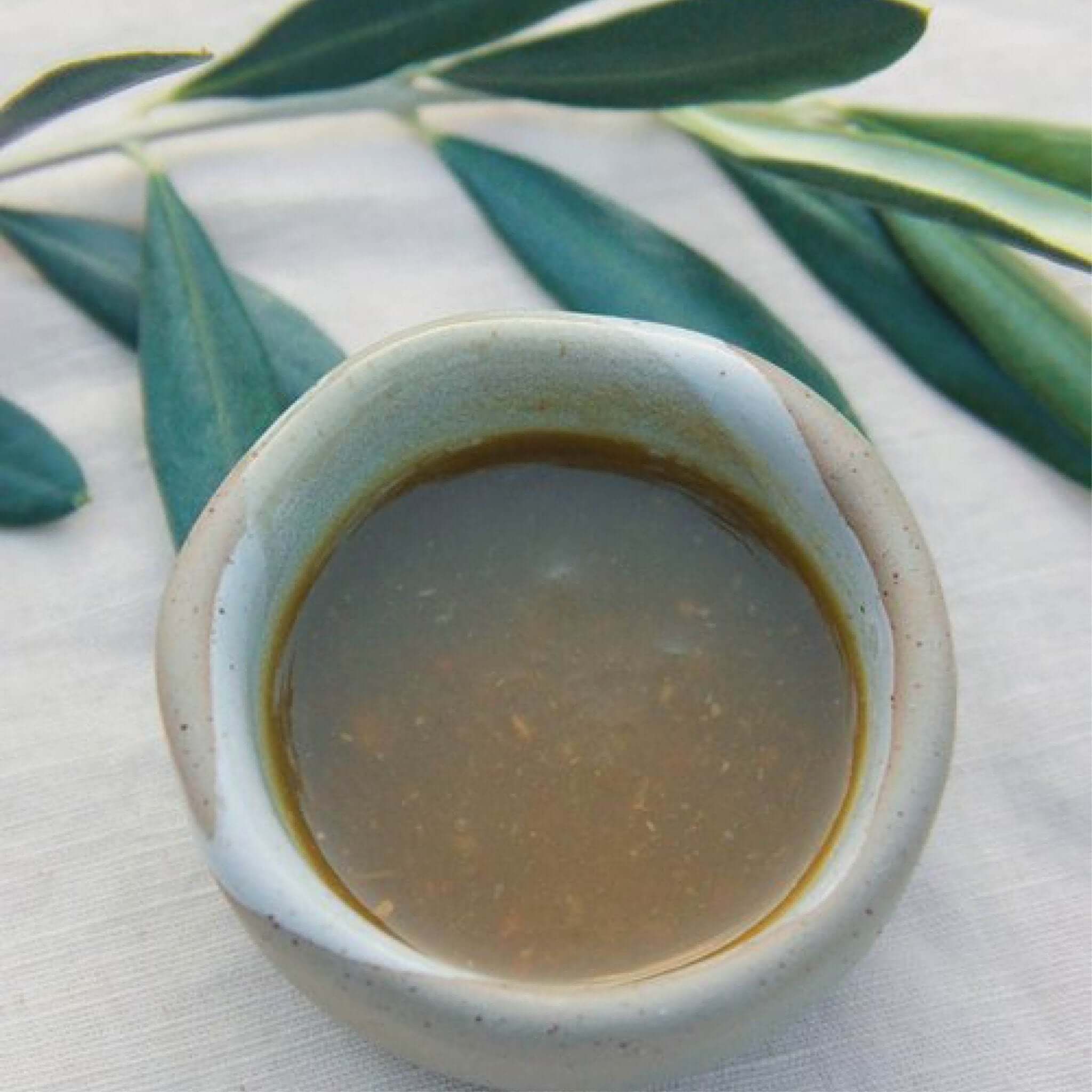 A rustic ceramic bowl filled with brownish liquid on a light-colored fabric surface. Green leaves, possibly olive, in the background. The liquid is Crack Fox Bintango hot sauce