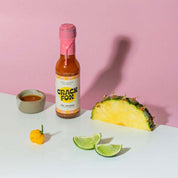 A Crack Fox hot sauce bottle labeled "Hot Jamaica" with a pink wax seal on a white surface against a pink background. Nearby: a yellow pepper, lime wedges, pineapple slice, and a bowl of sauce.