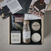 A curated gift box with various self-care items, including caramel milk sprinkle, natural tea soak, black clay facial soap, hand-poured soy candle, and nourishing hand cream.