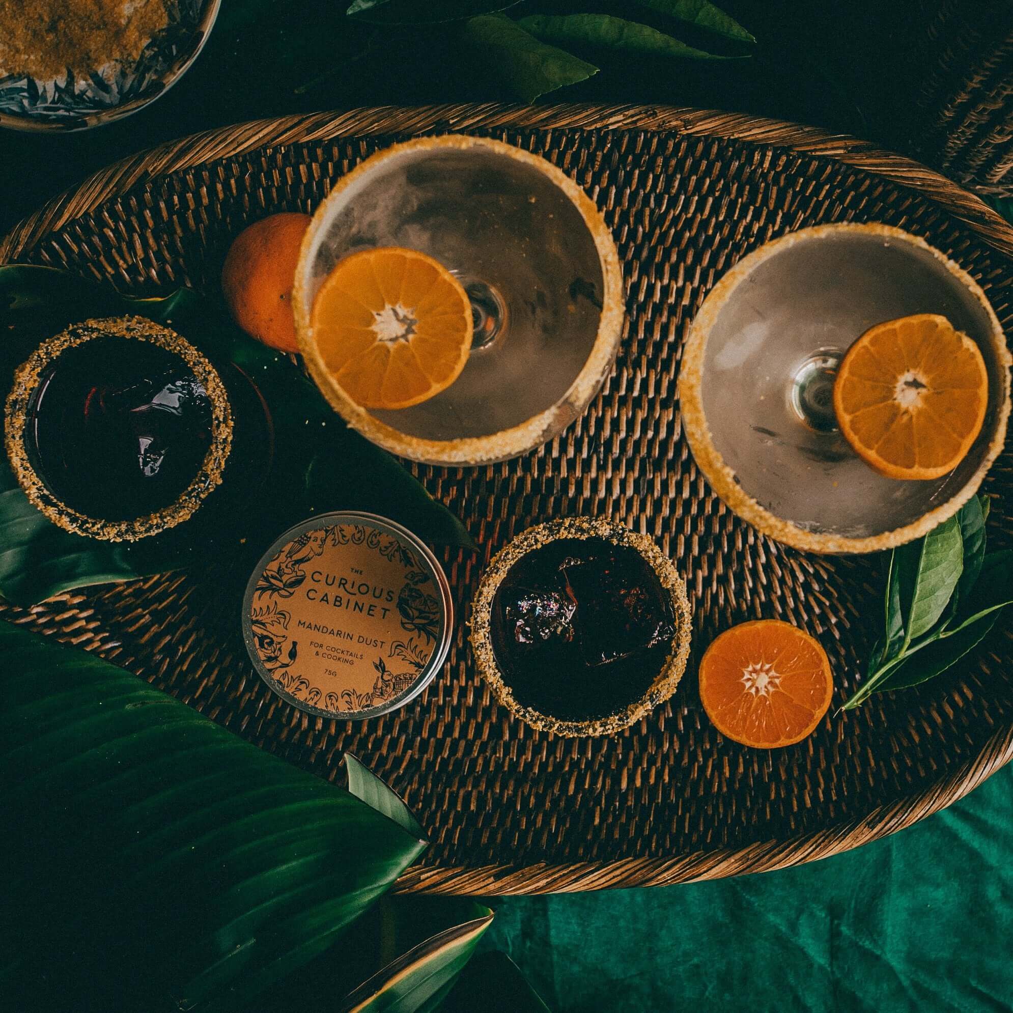 Wicker tray with two empty cocktail glasses with sugared rims, three smaller glasses with dark liquid, a tin labeled "The Curious Cabinet Mandarin Dust for Cocktails," green leaves, and half an orange.