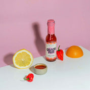 A bottle of "CRACK FOX" Citrusracha hot sauce surrounded by ingredients: a lemon half, orange half, habanero peppers, and a bowl of sauce. Pink backdrop.