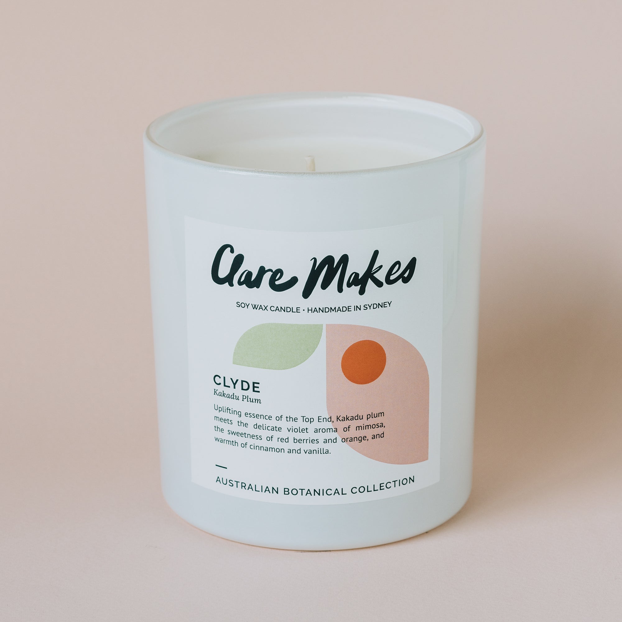 A white jar candle with a black lid. The label reads &quot;Clare Makes&quot; and &quot;Kakadu Plum.&quot; The candle is described as having an uplifting scent with notes of Kakadu plum, violet, mimosa, red berries, orange, cinnamon, and vanilla. It is part of the Australian 