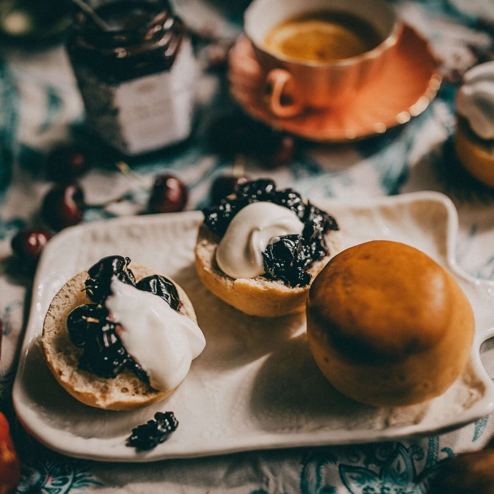 A white plate with a split bun topped with dark fruit preserve and cream. A jar of fruit preserve, scattered cherries, and an orange cup of coffee are in the background. The scene is on a patterned tablecloth.