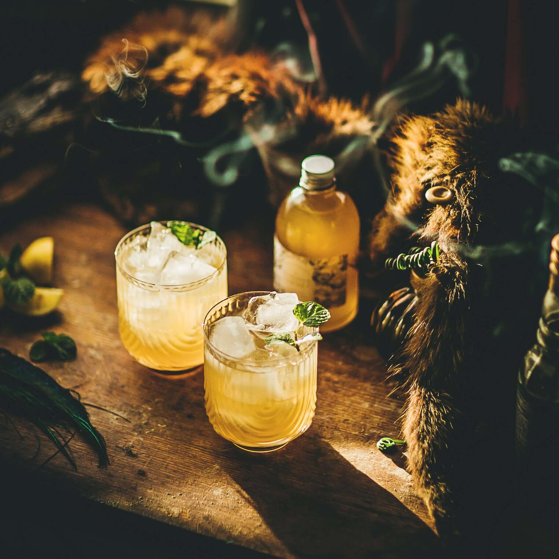 Two glasses of yellowish drink with ice and mint leaves on a wooden surface. Small bottle of yellow liquid, lemon wedges, mint, warmly lit setting.
