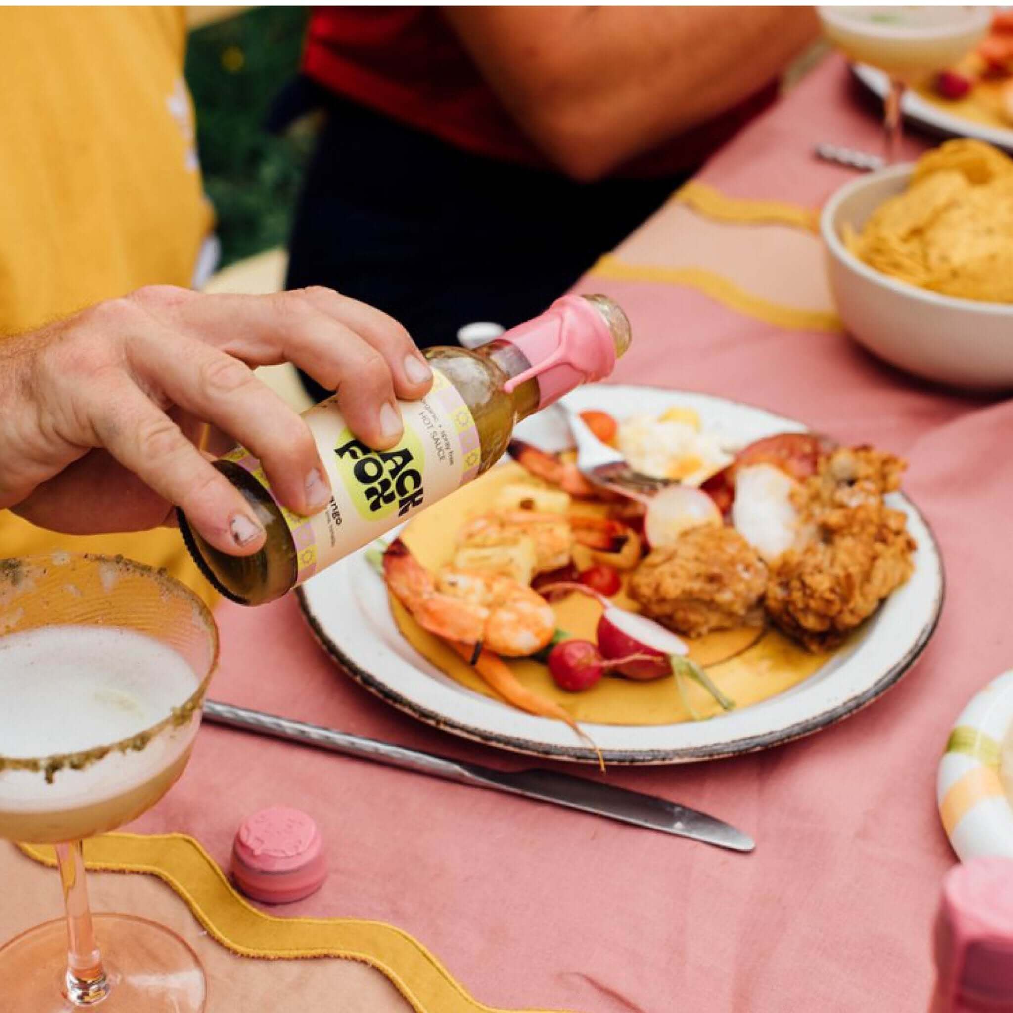 A close-up of a dining table with a plate of food and a hand holding a sauce bottle labeled "Jack Fruit." The plate contains shrimp, fried chicken, radishes, and a glass with a drink.