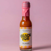 A 150 ml bottle of Crack Fox hot sauce with a pink wax seal on a pink background. Label reads "organic + spray free HOT SAUCE," "Hot Jamaica," and lists the ingredients.