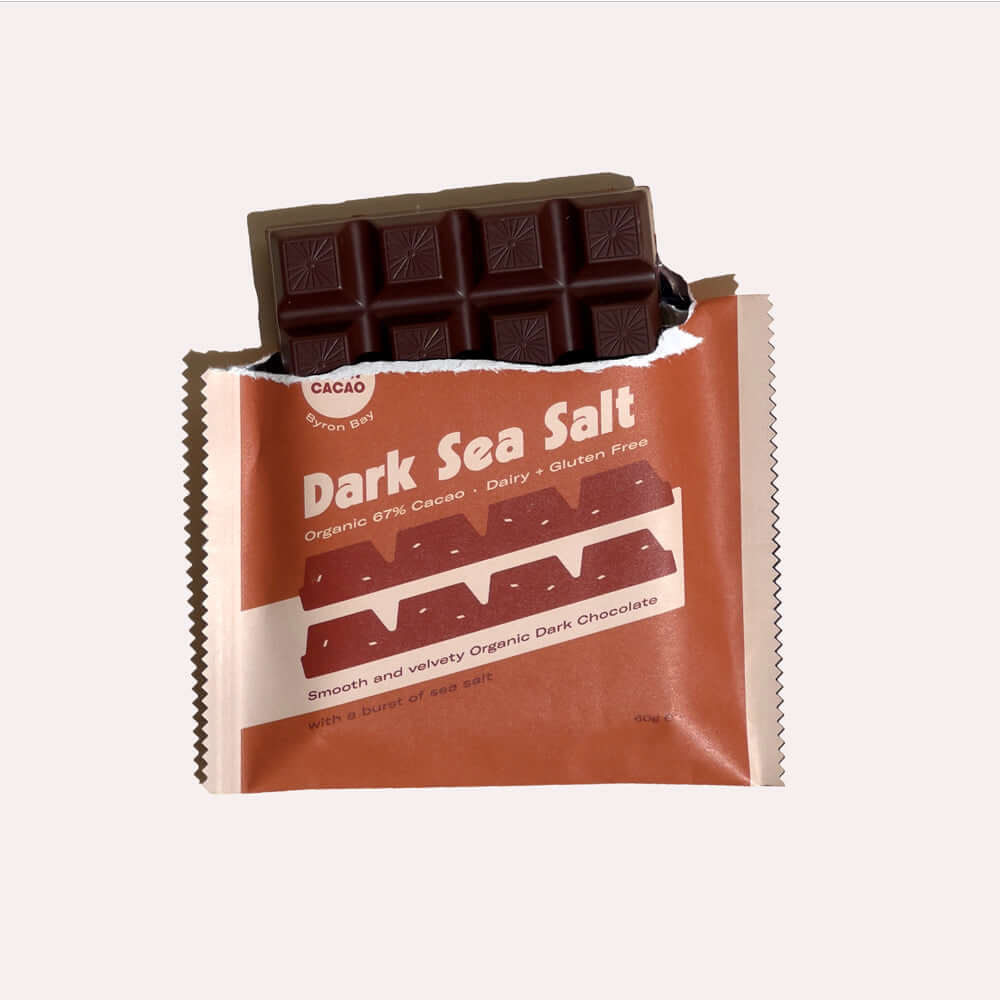 A close-up of a partially opened chocolate bar wrapper. The chocolate bar is dark brown with a visible snap, indicating it is a 67% cacao dark chocolate with sea salt. The wrapper is brown with white lettering that says "Byron Bay Cacao, Dark Sea Salt, Or