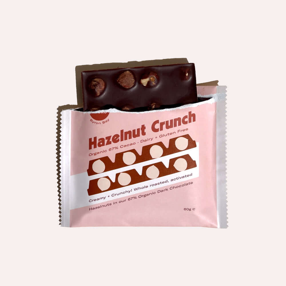 A close-up of an open chocolate bar wrapper. Inside, a dark chocolate bar is broken, revealing chunks of roasted hazelnuts. The wrapper is pink with white text that reads "Hazelnut Crunch, Organic 67% Cacao, Dairy & Gluten Free, Creamy + Crunchy Whole Roa