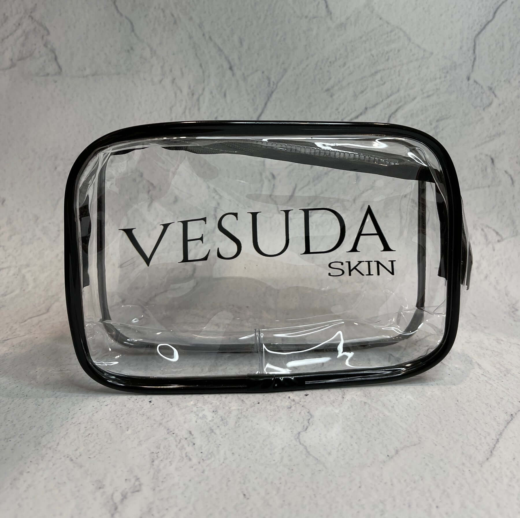 Clear cosmetic bag with black trim and "VESUDA SKIN" printed on the front. Background is a textured, light-colored surface.