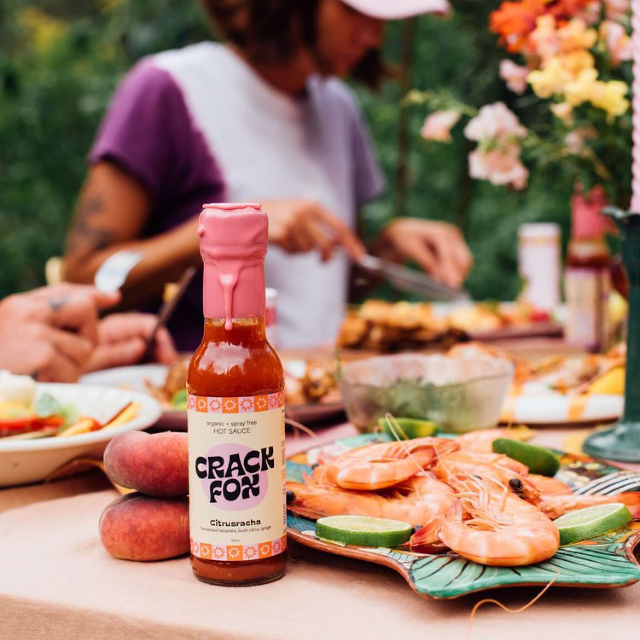 A bottle of "CRACK FOX" Citrusracha hot sauce on a table with prawns, lime slices, and peaches. Diners and vibrant flowers in the blurred background.