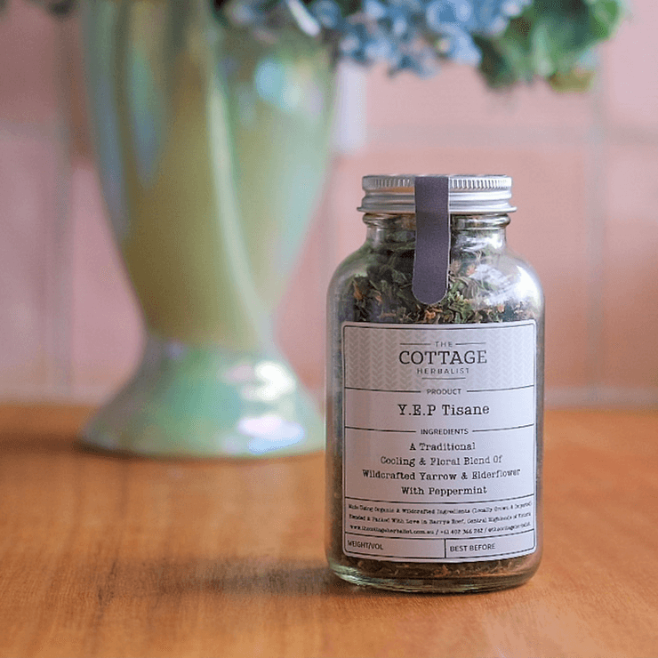 A glass jar filled with dried herbs sits on a wooden table. The label reads "The Cottage Herbalist Y.E.P Tisane" and lists the ingredients as a cooling blend of wildcrafted yarrow and elderflower with peppermint.