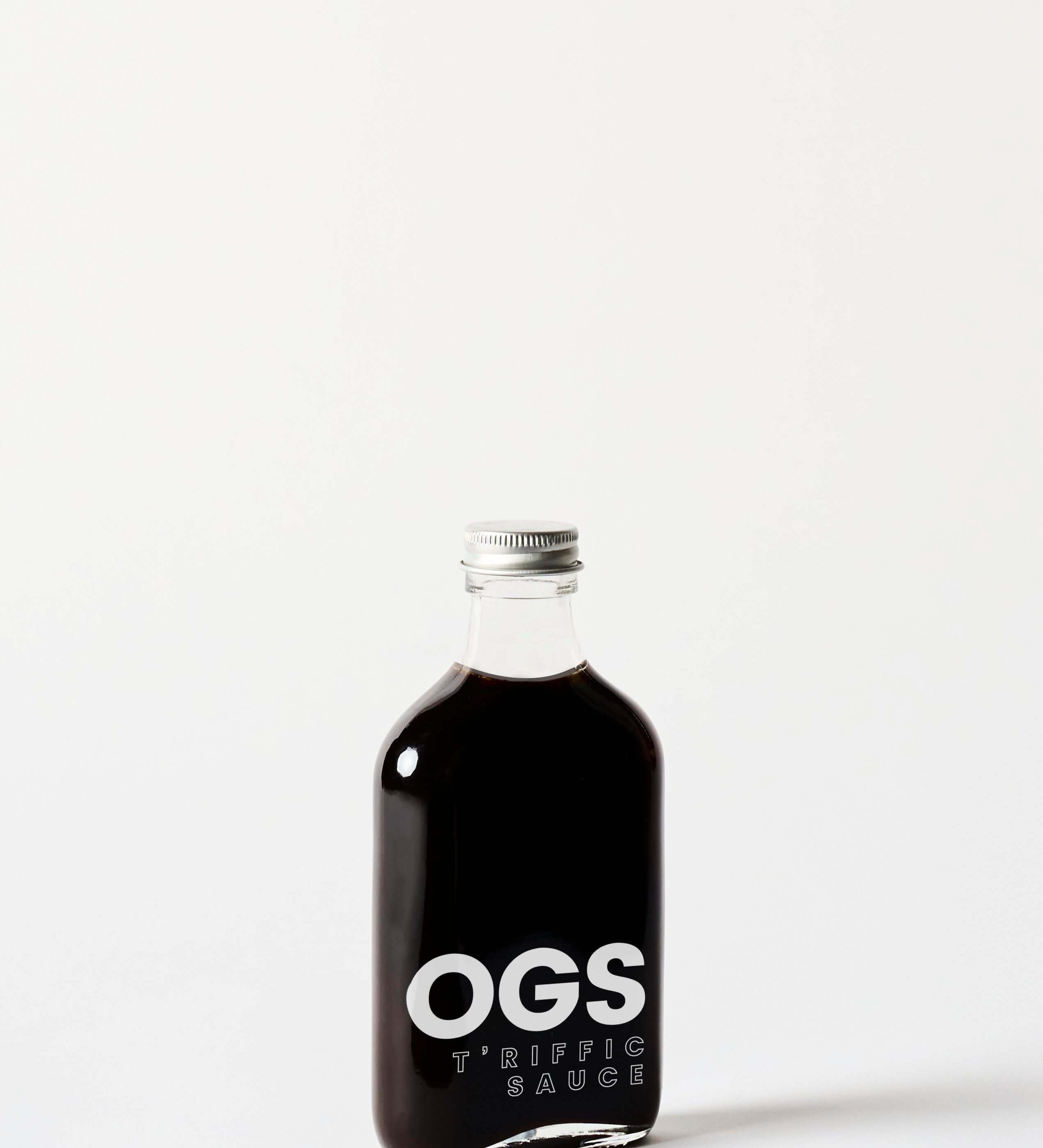 A glass bottle of OGS T'riffic Sauce. The sauce is a dark red color and the bottle has a silver cap. The label is white with black text. The bottle is standing upright on a white background.