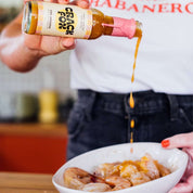 A person pours Crack Sauce "Hot Jamaica" from a bottle onto a bowl of raw shrimp. The person wears a white shirt with red text and dark jeans. The sauce is orange.