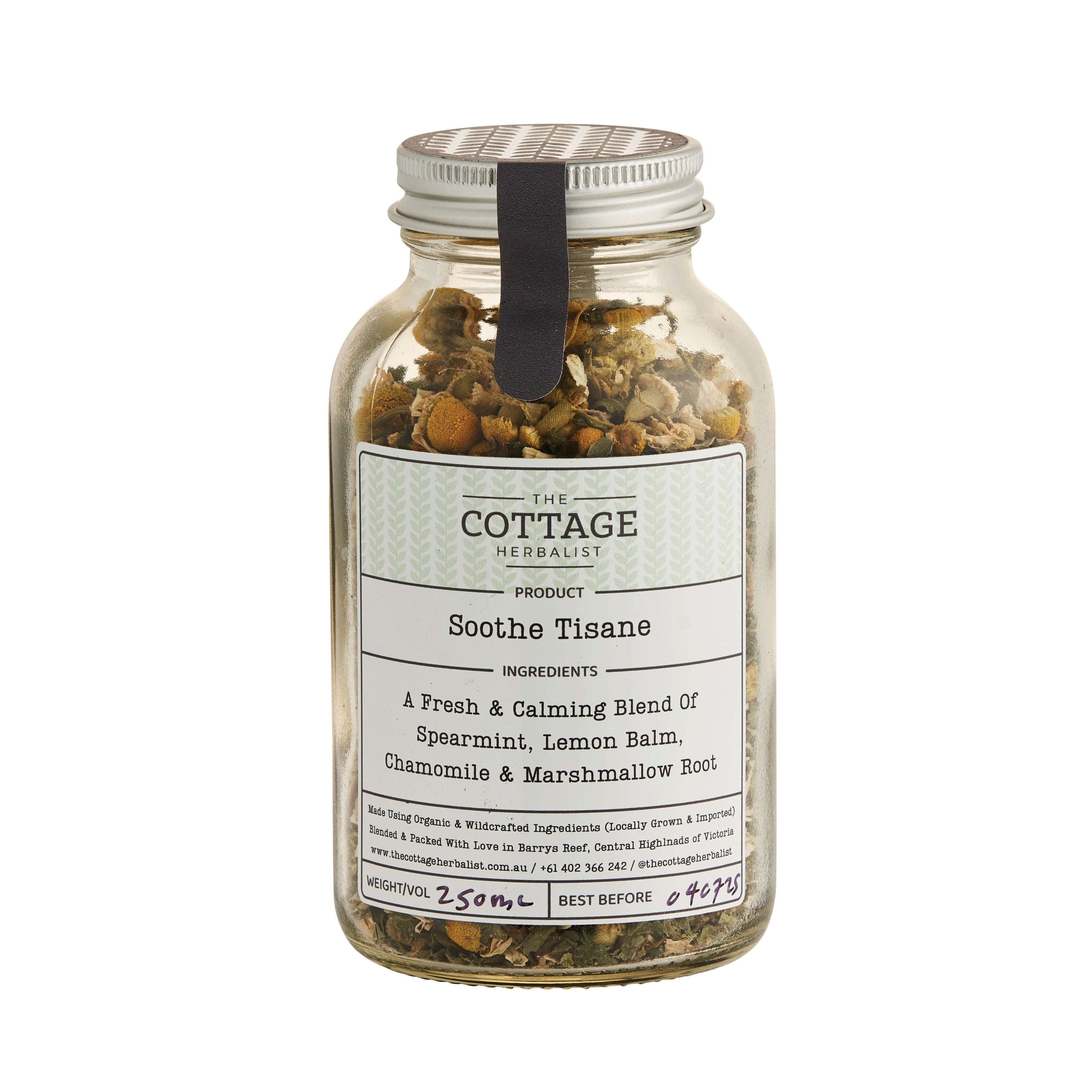 A glass jar filled with dried herbs sits on a wooden table. The label reads "The Cottage Herbalist Soothe Tisane" and lists the ingredients as a fresh & calming blend of spearmint, lemon balm, chamomile, and marshmallow root.