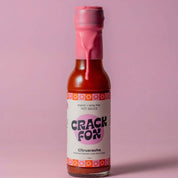 A bottle of "CRACK FOX" Citrusracha hot sauce with a pink wax seal, featuring fermented habanero, bush citrus, and ginger. Vibrant design on a pink backdrop.