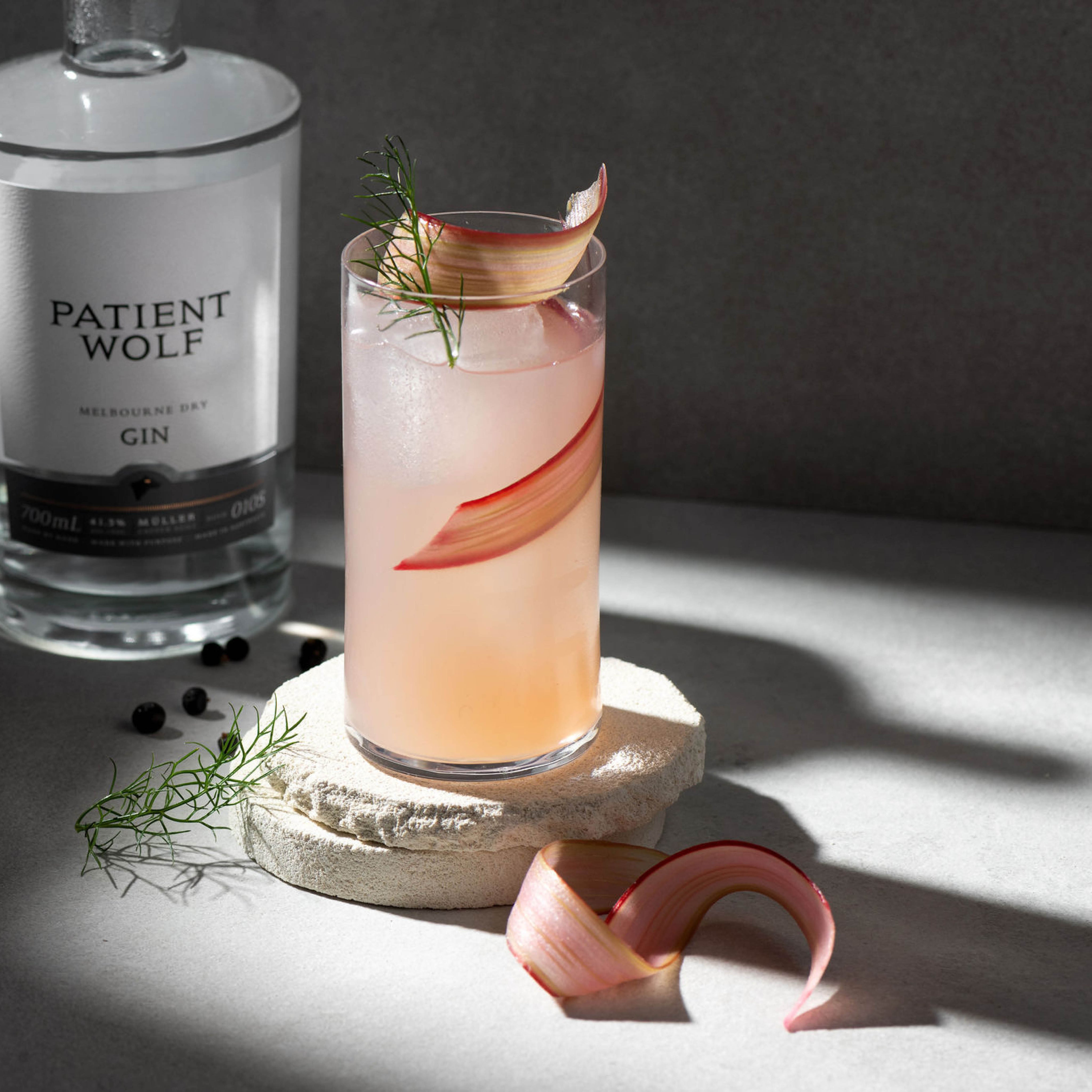 A light pink gin cocktail garnished with rhubarb and dill next to a bottle of Patient Wolf Melbourne Dry Gin. On a round stone coaster with juniper berries and rhubarb pieces around it.