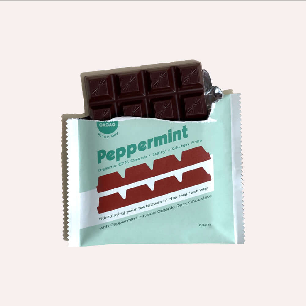 A close-up of an open chocolate bar wrapper. Inside, a dark chocolate bar is broken, revealing the peppermint infused chocolate. The wrapper is teal with white text that reads "Peppermint, Organic 67% Cacao, Dairy & Gluten Free, Stimulating your tastebuds