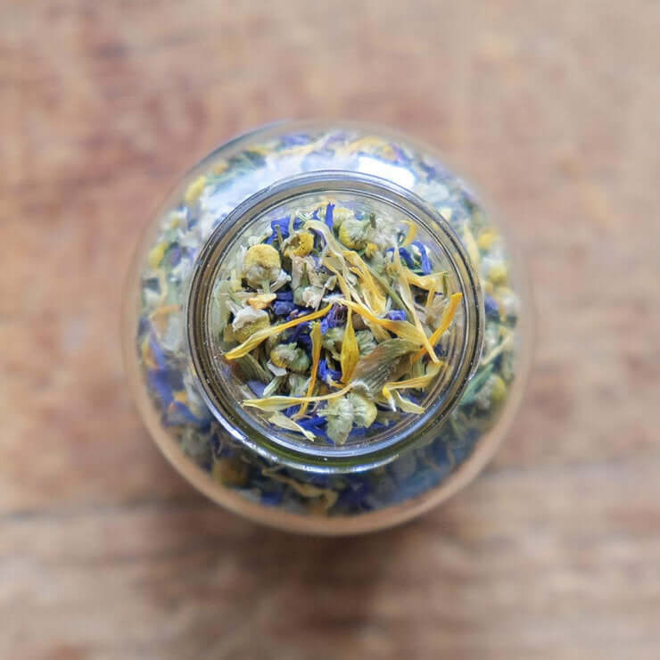 A top-down view of a glass jar filled with a colorful blend of dried herbs and flowers, including blue cornflowers, yellow chamomile, and other botanicals. The image evokes a sense of natural wellness and relaxation.