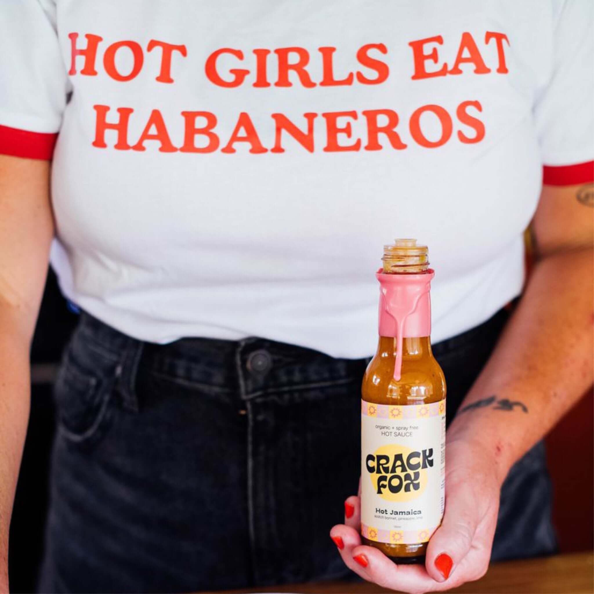 A person in a white t-shirt with "HOT GIRLS EAT HABANEROS" in red text, holding a bottle of Crack Fox Hot Jamaica sauce with a pink wax seal, outdoors.