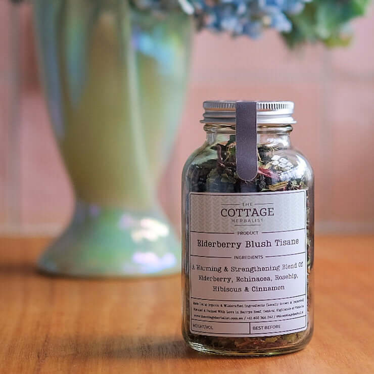 A glass jar filled with dried herbs sits on a wooden table, with a blurred vase of flowers in the background. The label reads "The Cottage Herbalist Elderberry Blush Tisane" and lists the ingredients as a warming & strengthening blend of elderberry, echin