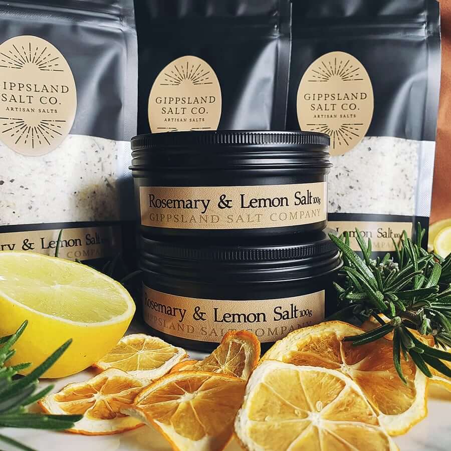 A collection of Gippsland Salt Co. artisan salts. Black tins and pouches of salt are displayed alongside fresh lemons, rosemary sprigs, and dried citrus slices. The image highlights the brand's focus on natural ingredients and flavors.