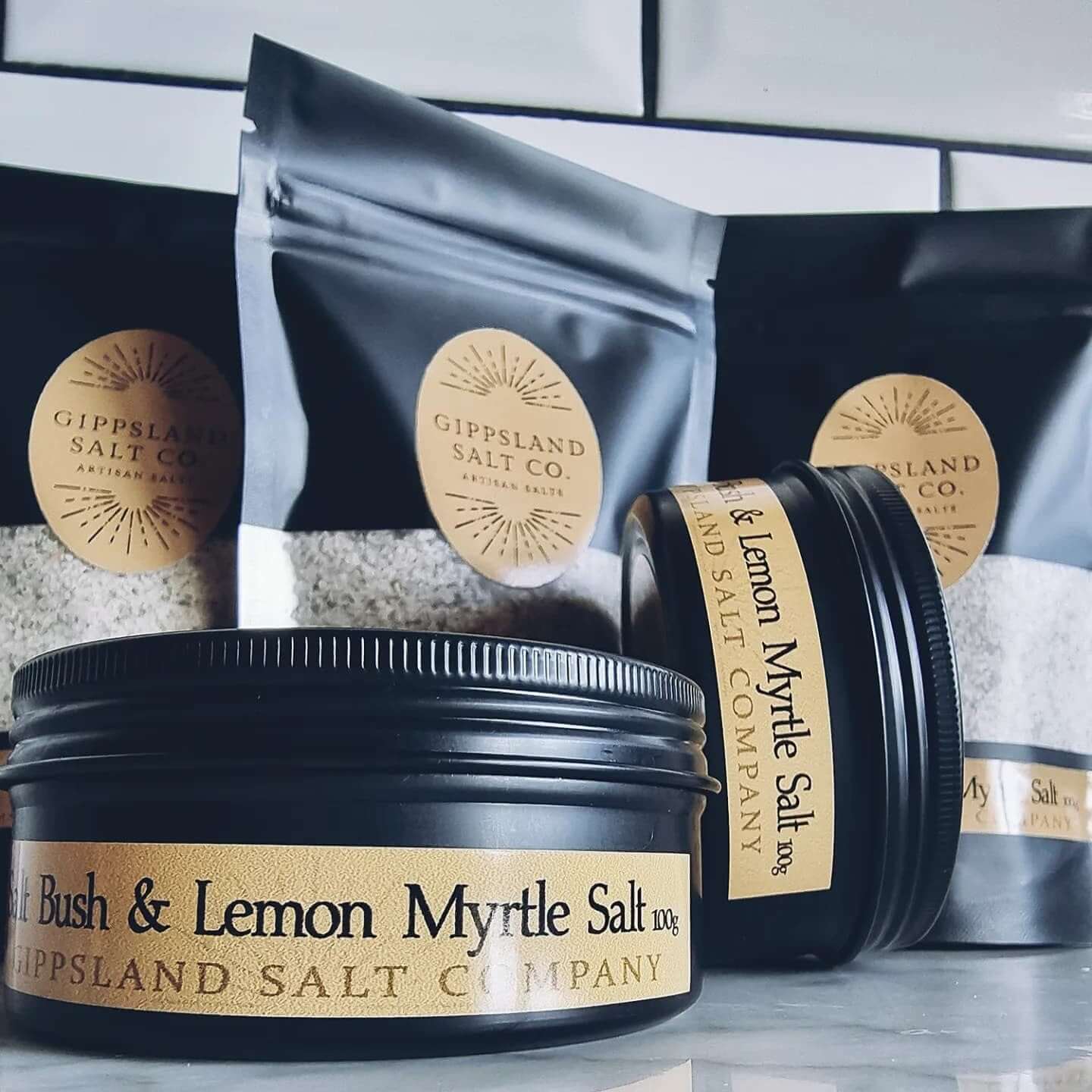 A collection of Gippsland Salt Co. artisan salts. Black tins and pouches of salt are displayed on a counter, showcasing flavors like Salt Bush & Lemon Myrtle. The image highlights the brand's variety and focus on natural ingredients.