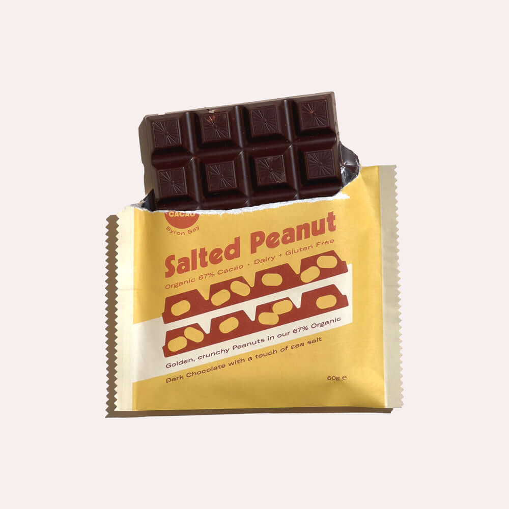 A close-up of an open chocolate bar wrapper. Inside, a dark chocolate bar is broken, revealing the salted peanuts. The wrapper is yellow with white text that reads "Salted Peanut, Organic 67% Cacao, Dairy & Gluten Free, Golden, crunchy Peanuts in our 67% 