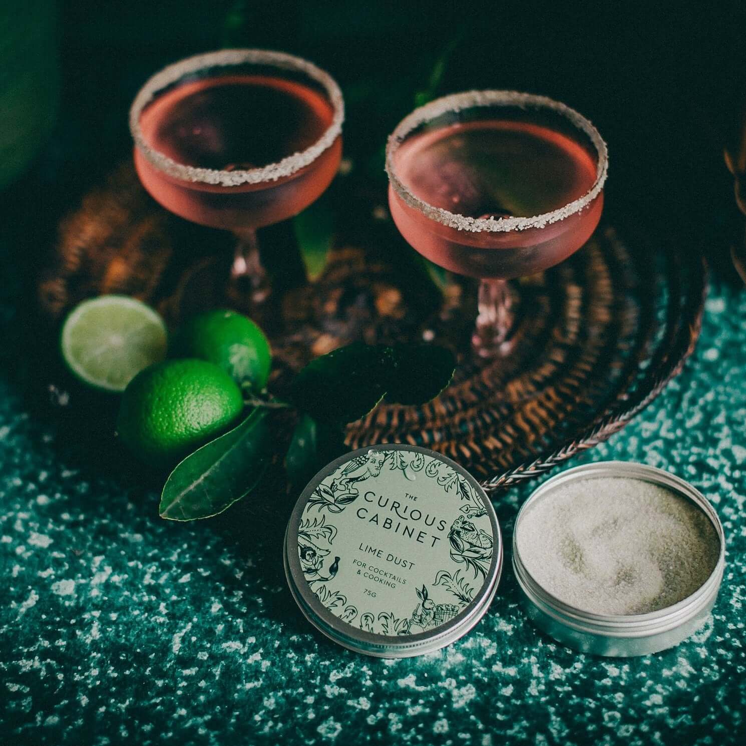 Two cocktail glasses with pinkish-red drinks and sugared rims on a woven tray. Three whole limes, one halved, and an open tin of "Lime Dust" beside it.