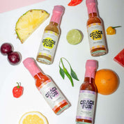 Four Crack Fox hot sauce bottles with pink caps on a white surface, surrounded by fruits and vegetables like pineapple, lime, plums, peppers, and citrus slices.