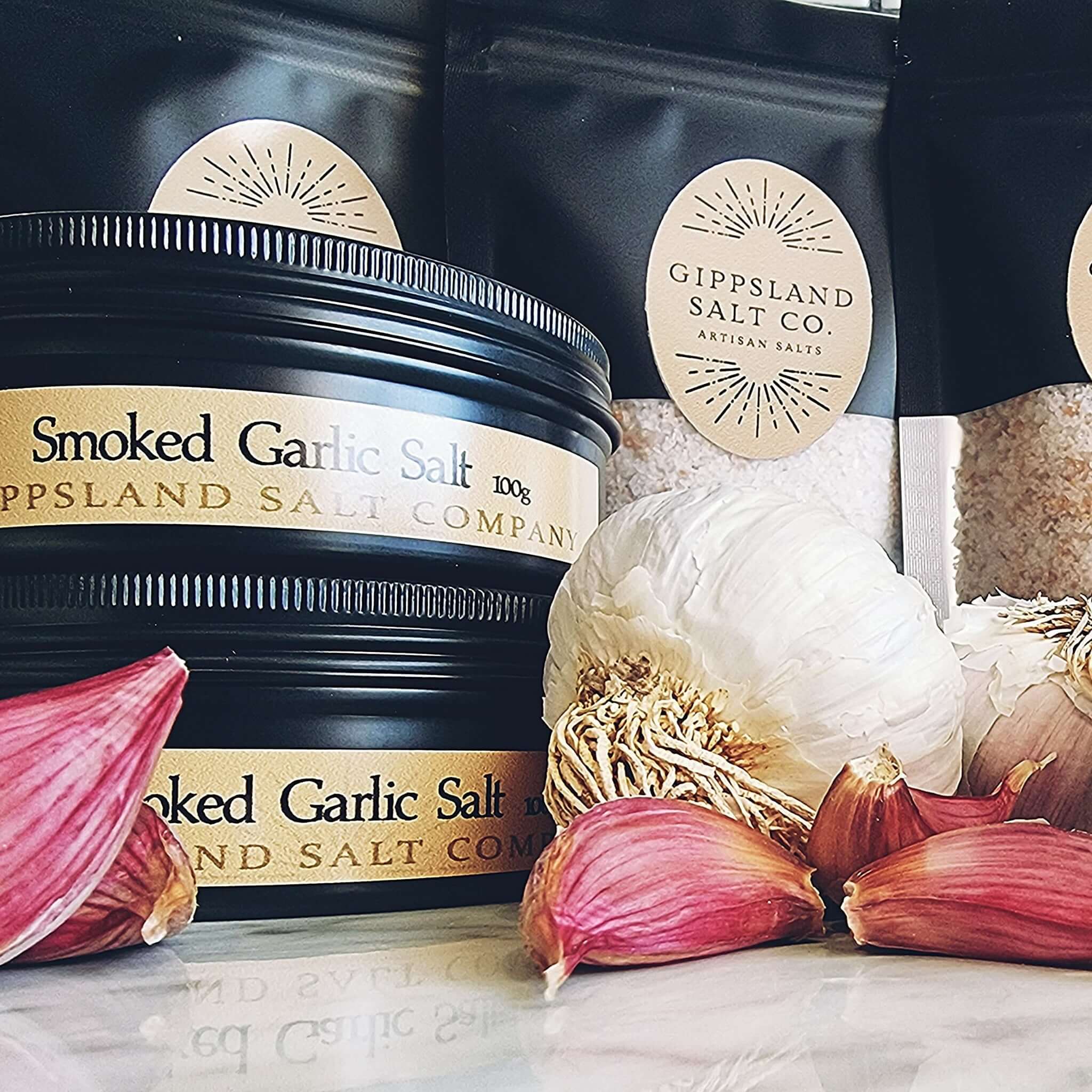 A black tin of Gippsland Salt Co. Smoked Garlic Salt sits on a counter next to several bulbs of garlic. The image emphasizes the connection between the salt and its key ingredient. The label design is simple and elegant.
