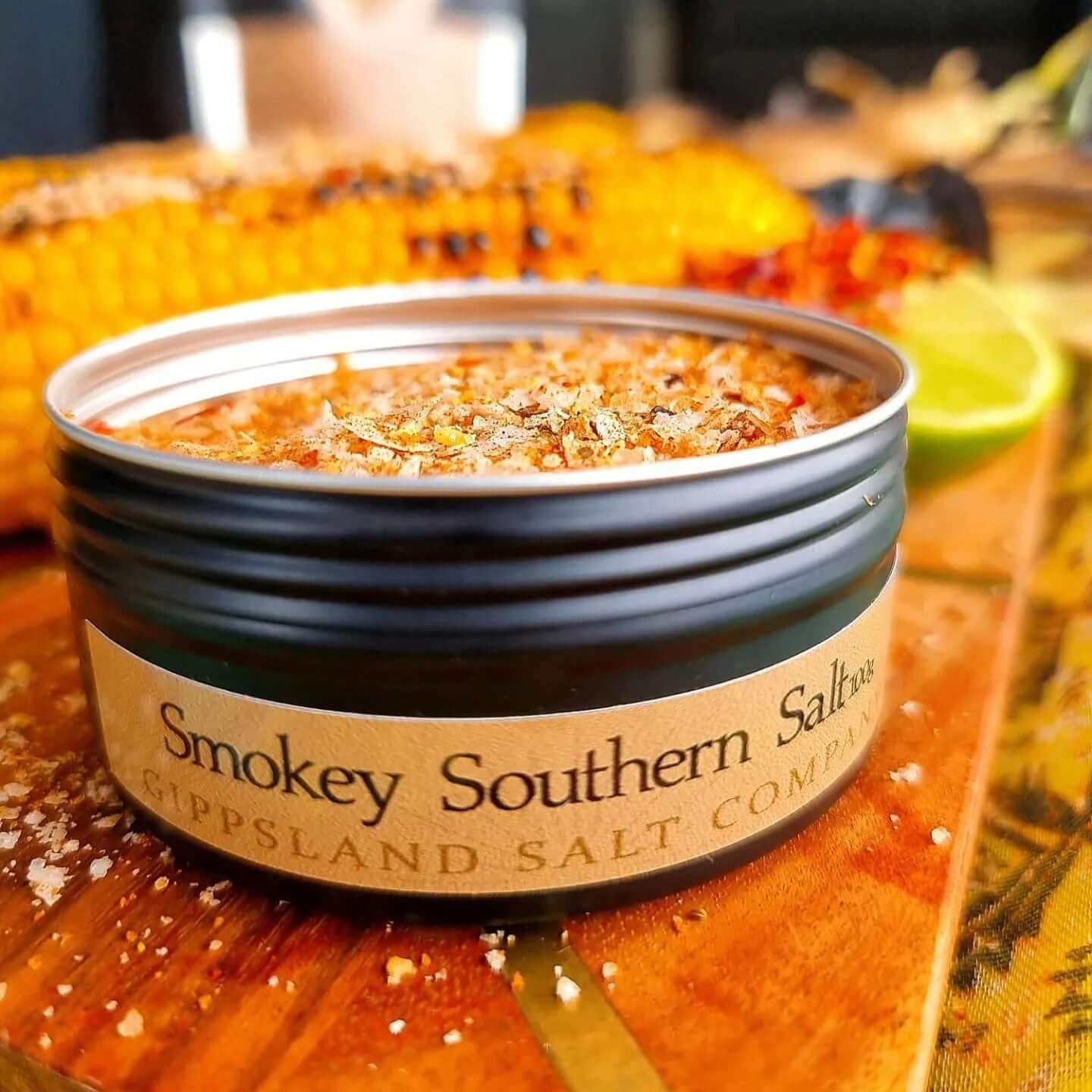 A close-up of an open tin of Gippsland Salt Company's Smokey Southern Salt. The salt has a reddish hue, suggesting the presence of chili flakes. A grilled corncob is visible in the background, hinting at the salt's intended use.