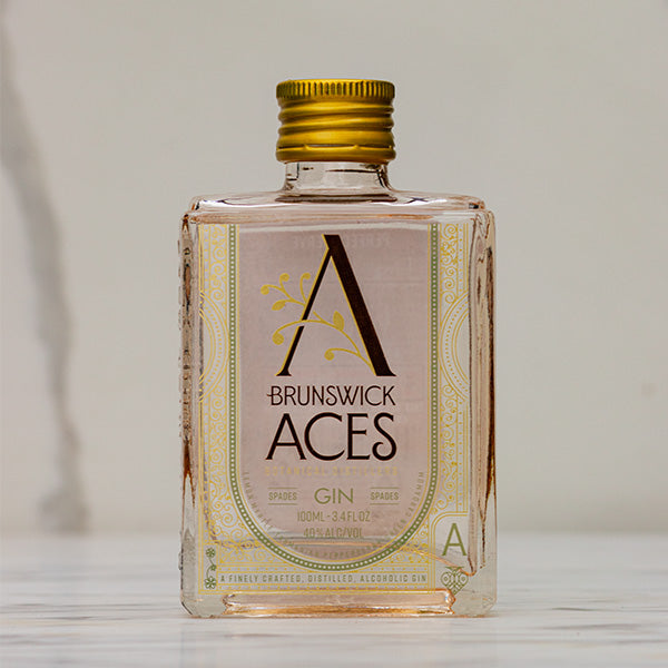 A miniature square bottle of Brunswick Aces Spades Gin with a gold cap. The label features an ornate "A" logo and highlights the use of premium botanicals. The gin is 100ml with an alcohol content of 40%.