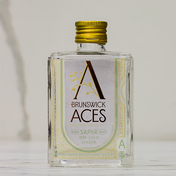 A miniature square bottle of Brunswick Aces Sapiir, an alcohol-free distilled botanical spirit. The label features an ornate "A" logo and highlights the use of native Australian desert lime and mango. The bottle is 100ml and 0% alcohol.