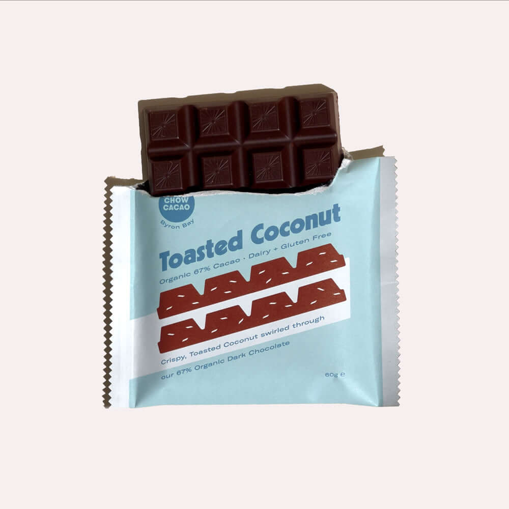 A close-up of an open chocolate bar wrapper. Inside, a dark chocolate bar is broken, revealing the toasted coconut pieces. The wrapper is light blue with white text that reads "Toasted Coconut, Organic 67% Cacao, Dairy & Gluten Free, Crispy, Toasted Cocon