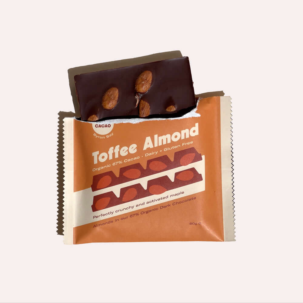 A close-up of an open chocolate bar wrapper. Inside, a dark chocolate bar is broken, revealing the toffee and almond pieces. The wrapper is orange with white text that reads "Toffee Almond, Organic 67% Cacao, Dairy & Gluten Free, Perfectly crunchy and act