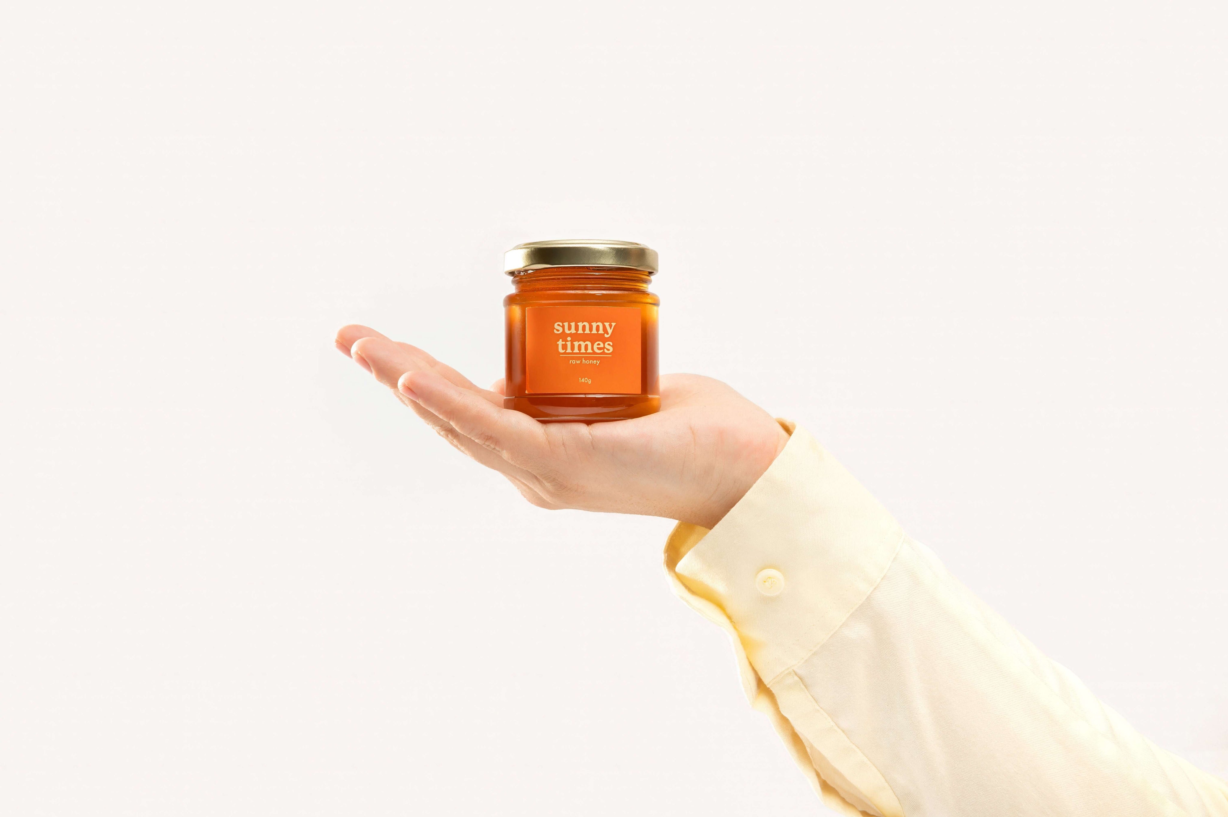 A hand holding a jar of Sunny Times Raw Honey against a plain white background. The honey jar has an orange label with white text. A yellow sleeve cuff is visible on the right.