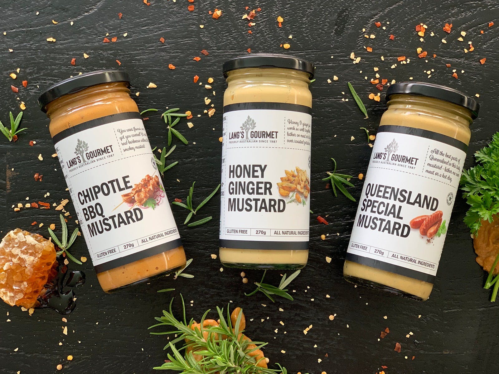 A vibrant product shot of Lang's Gourmet mustards: Chipotle BBQ, Honey Ginger, and Queensland Special. Displayed on a dark background with fresh rosemary, honeycomb, and spices scattered around, emphasising their all-natural and flavourful appeal.