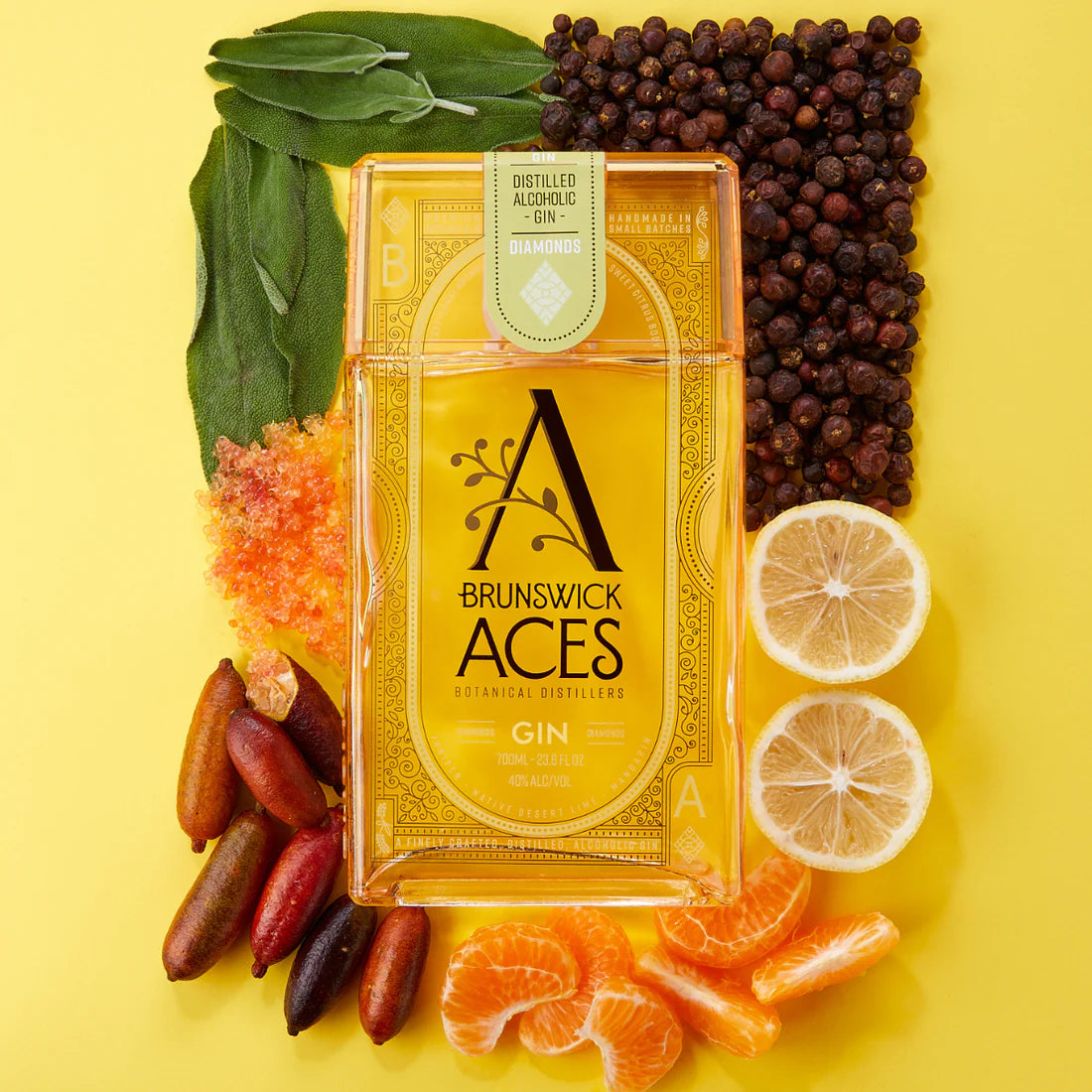 A bottle of Brunswick Aces Gin is surrounded by botanicals, including lemons, mandarins, juniper berries, sage leaves, and other exotic ingredients. The bottle design features a stylized "A" and ornate details, highlighting the gin's handcrafted nature.