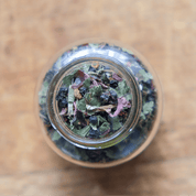 A top-down view of a glass jar filled with a blend of dried herbs and berries, including dark purple elderberries, rosehips, and possibly cinnamon sticks. The image evokes a sense of warmth and wellness.