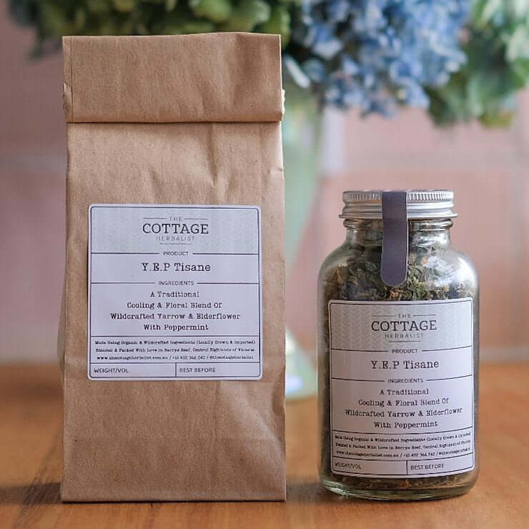 Two glass jars filled with dried herbs sit on a wooden table. The labels read "The Cottage Herbalist Y.E.P Tisane" and list the ingredients as a cooling blend of wildcrafted yarrow and elderflower with peppermint.