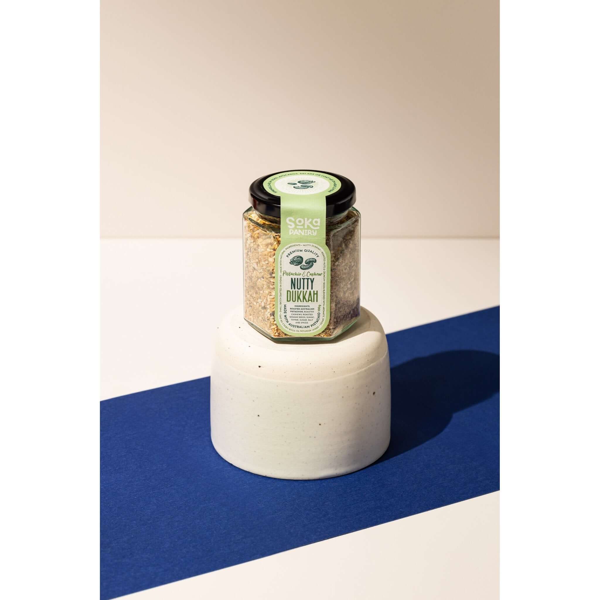 A jar of Soka Pantry Nutty Dukkah sits on a white, textured cylindrical stand against a split white and dark blue background. The jar has a black lid and a green label with white and black text. The dukkah inside is a coarse blend of nuts and spices.