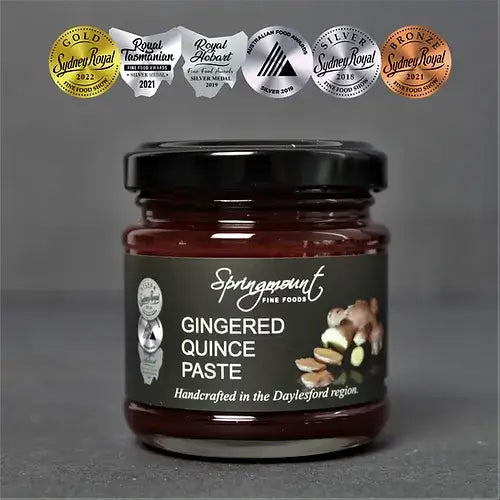 A jar of Springmount Gingered Quince Paste sits against a dark grey background. It has a black lid and a label with award stickers. The label shows an illustration of a sliced quince and ginger. The paste is a deep reddish-brown.