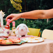 A white rabbit sitting at a meal table with various items, including sauce bottles, a candle, and a person's arm holding a bottle labeled "CRAZY FOODS" outdoors.