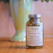 A glass jar filled with dried herbs sits on a wooden table. The label reads "The Cottage Herbalist Sweet Dreams Tisane" and lists the ingredients as a relaxing blend of chamomile, skullcap, passionflower, hops, cornflowers, calendula, and lavender.