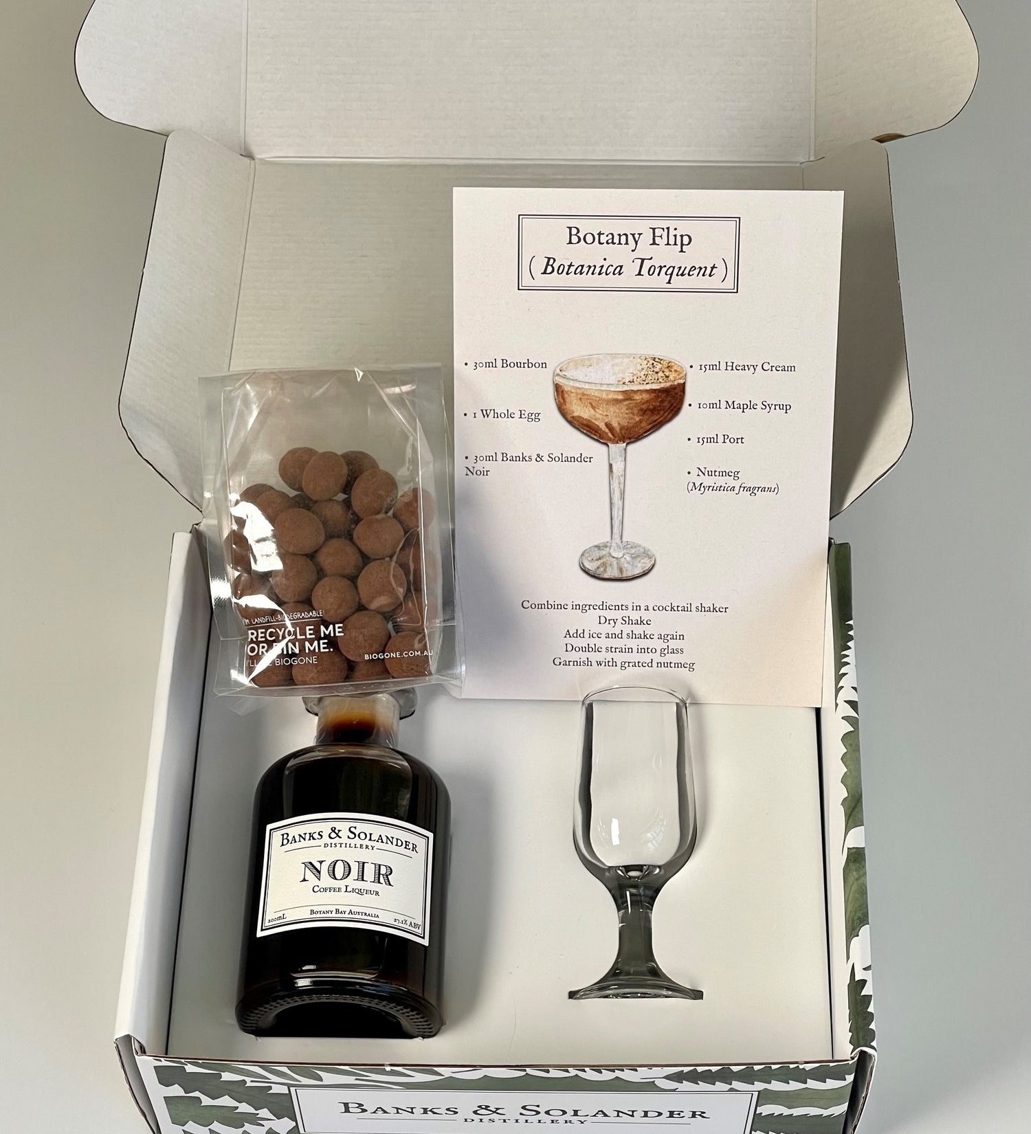 A gift box containing all the ingredients to make a "Botany Flip" cocktail. Inside are miniature bottles of Banks & Solander Noir liqueur, a recipe card, a small glass, and a bag of chocolate almonds.